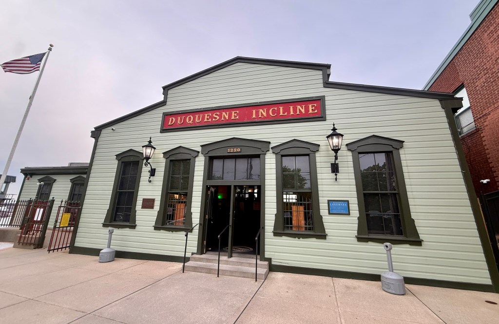 Exterior view of the Duquesne Incline building, featuring a green wooden facade and a prominent red sign, with an American flag in the background.