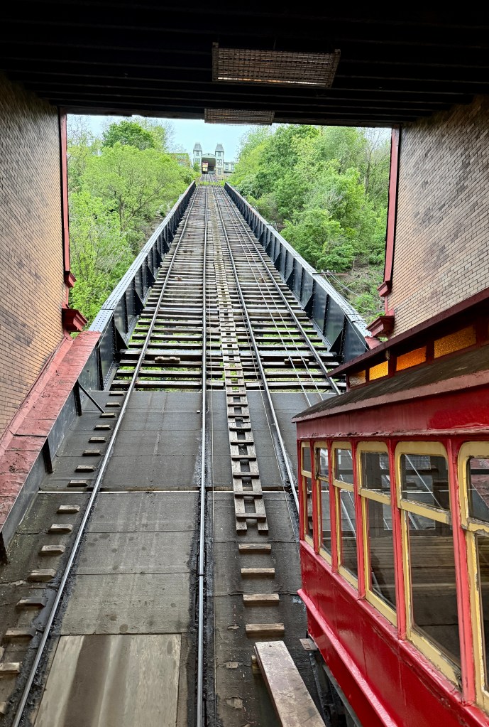 View from inside a funicular railway station, featuring a steep track leading uphill with a red train car on the right and greenery in the background.