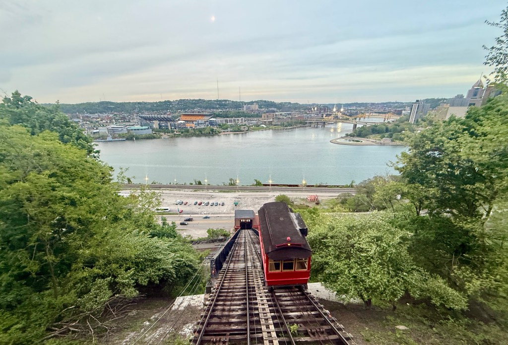 A view from above featuring a red incline train descending towards a river, surrounded by green trees and a city skyline in the background.