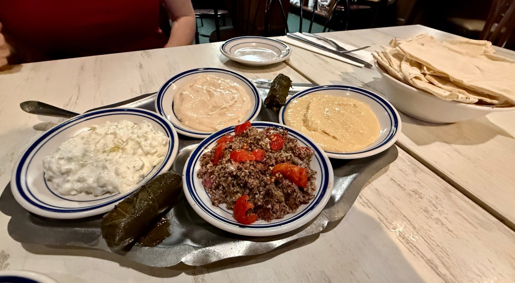 A platter with four small dishes of various dips, including a creamy spread, a hummus-like dip, a yogurt-based dish, and a spiced grain salad with tomatoes, accompanied by stuffed grape leaves and a bowl of flatbread.