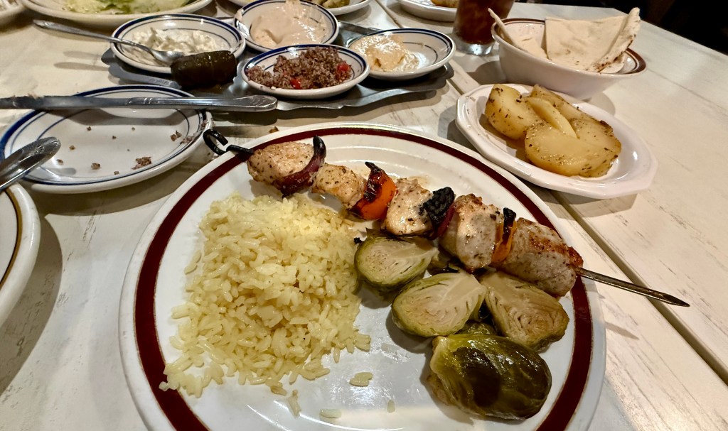 A plate featuring grilled chicken skewers with bell peppers, served with rice and Brussels sprouts. In the background, several side dishes and pita bread can be seen.