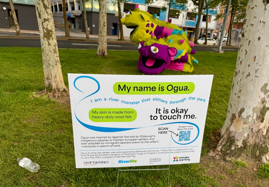 A colorful sculpture named Ogua, representing a river monster, with a sign describing it as a friendly character that slithers through the park. The sign encourages people to touch it and provides information about the artist and its inspiration.