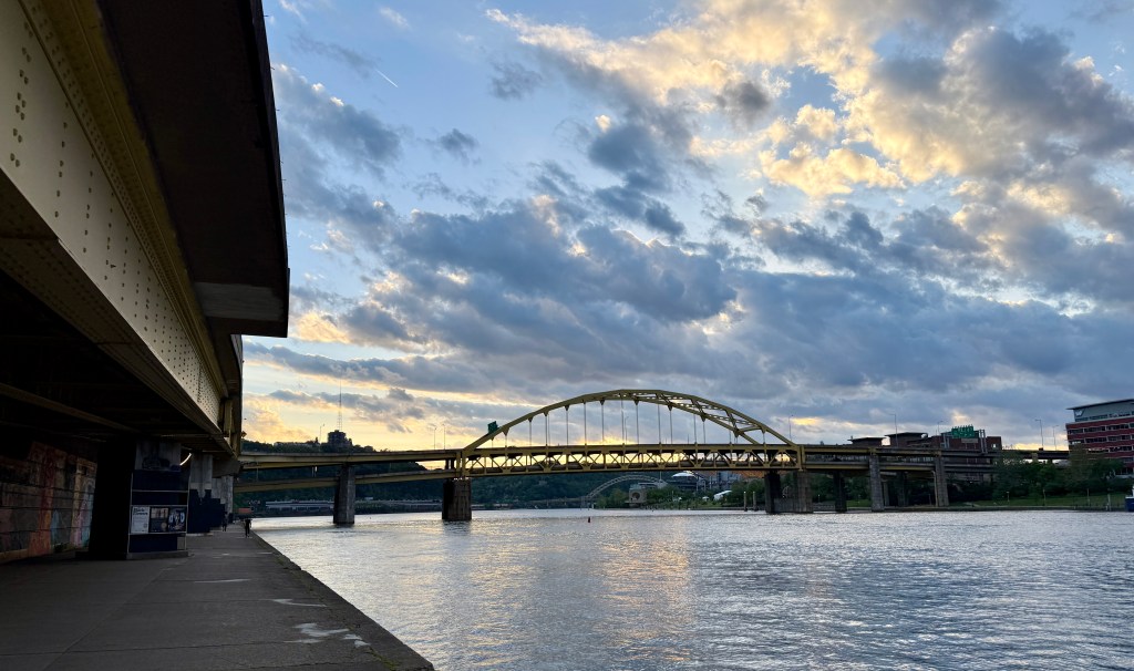 View of a river with a bridge in the background under a cloudy sky during sunset, featuring reflections on the water.