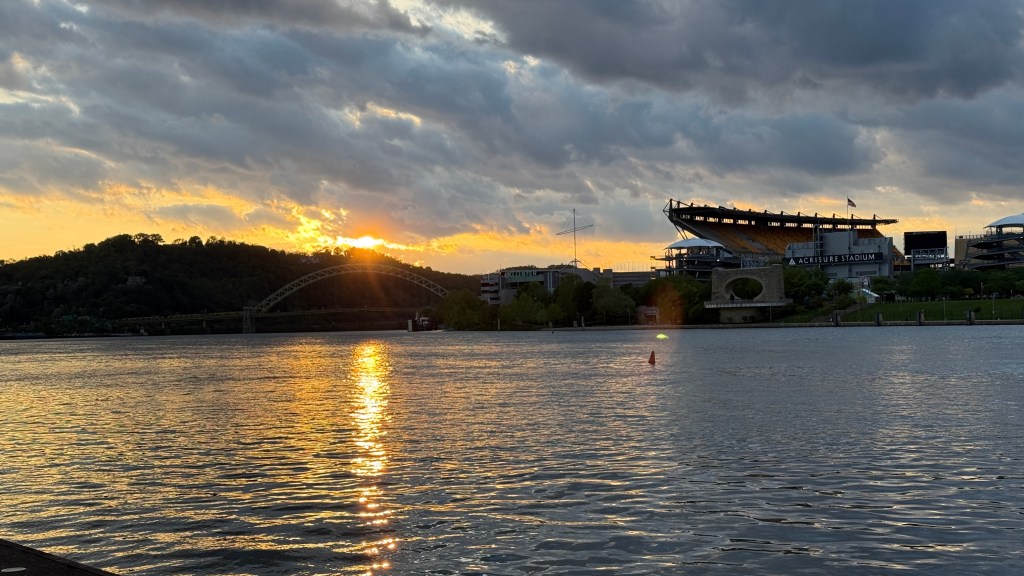 A sunset view over a river with reflections on the water, featuring a bridge in the background and a large stadium on the right.