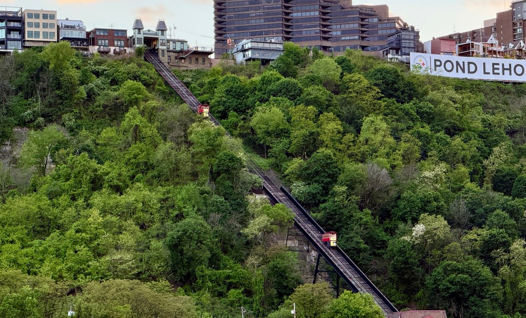View of a hillside featuring a funicular railway track ascending through lush greenery, with buildings in the background, and a large advertisement sign.