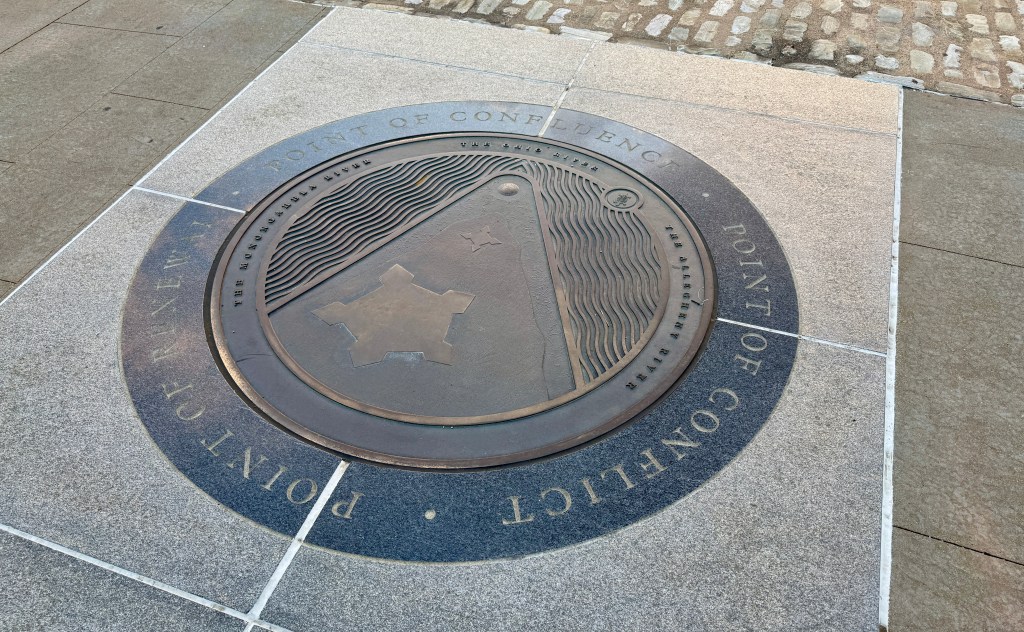 A bronze seal embedded in the ground, depicting the 'Point of Confluence' with stylized representations of land and water, marked by the surrounding text.