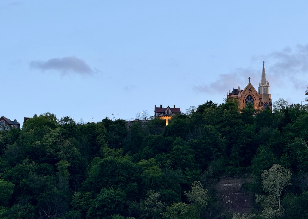 Hilly landscape with lush green trees and a Gothic-style church tower in the background during twilight.
