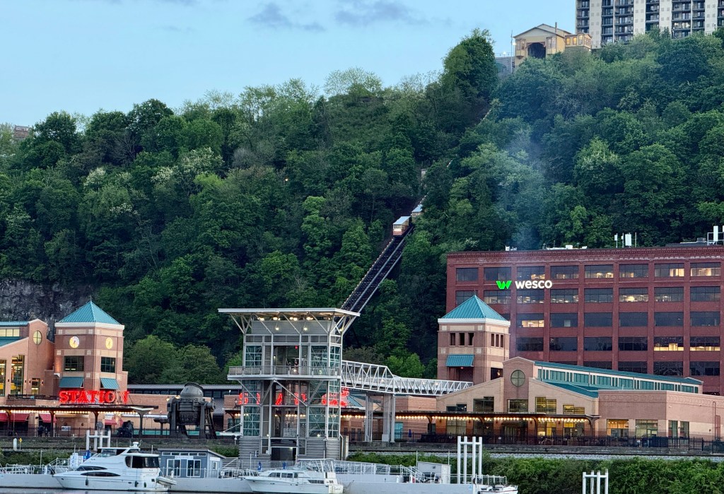 A view of a hillside with a funicular railway, shops, and buildings at the base, surrounded by dense greenery.