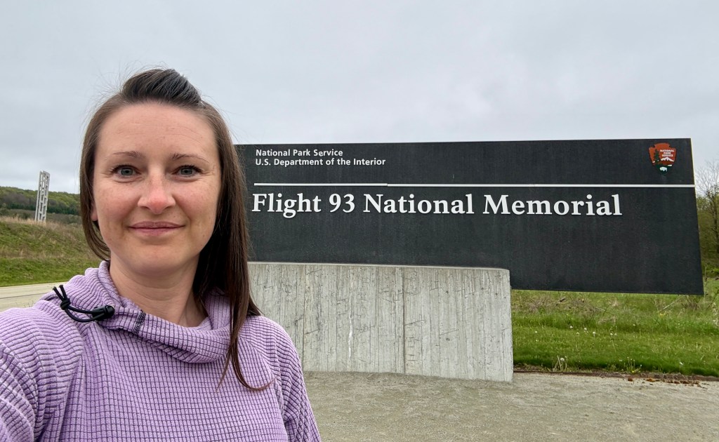 A woman stands in front of the Flight 93 National Memorial sign, with green grass and a cloudy sky in the background.
