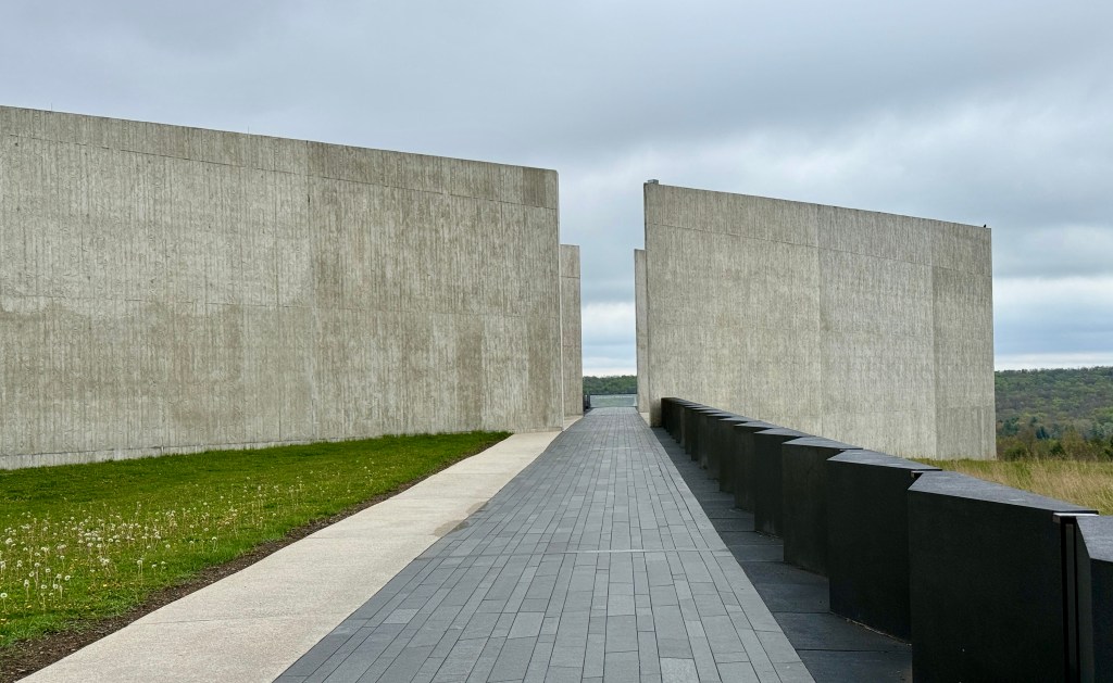 A pathway leads between two large concrete walls with a grassy area on one side, under a cloudy sky.