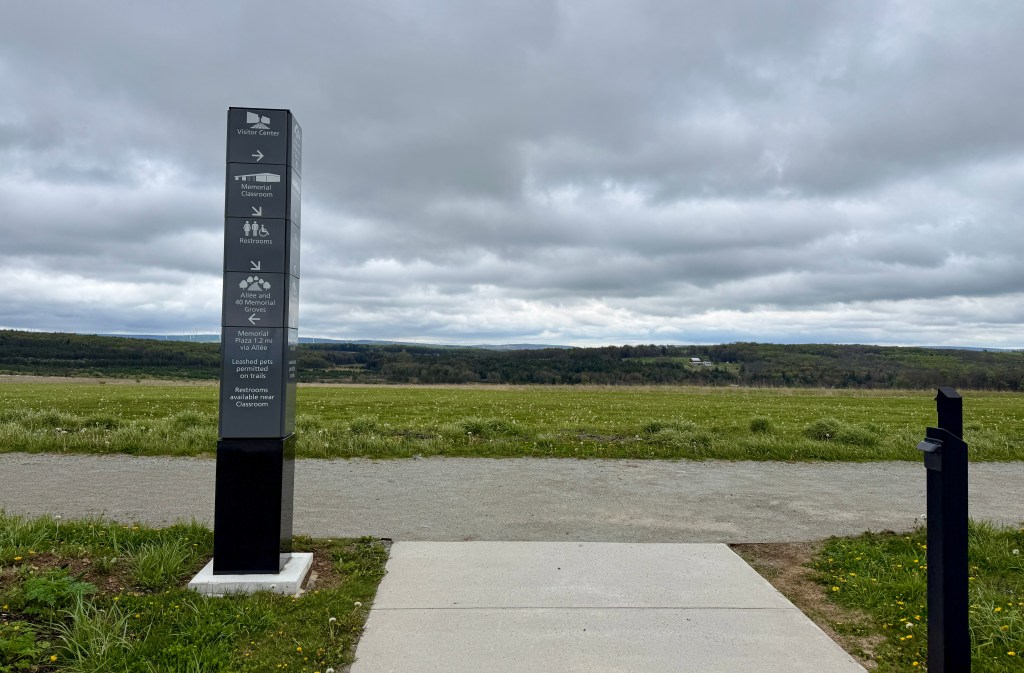 Signage at an outdoor area displaying directions and information for the Visitor Center, Memorial Classroom, restrooms, and trails, with a cloudy sky and green landscape in the background.