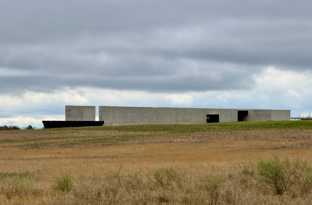 A modern, minimalist concrete building set against a cloudy sky and grassy landscape.