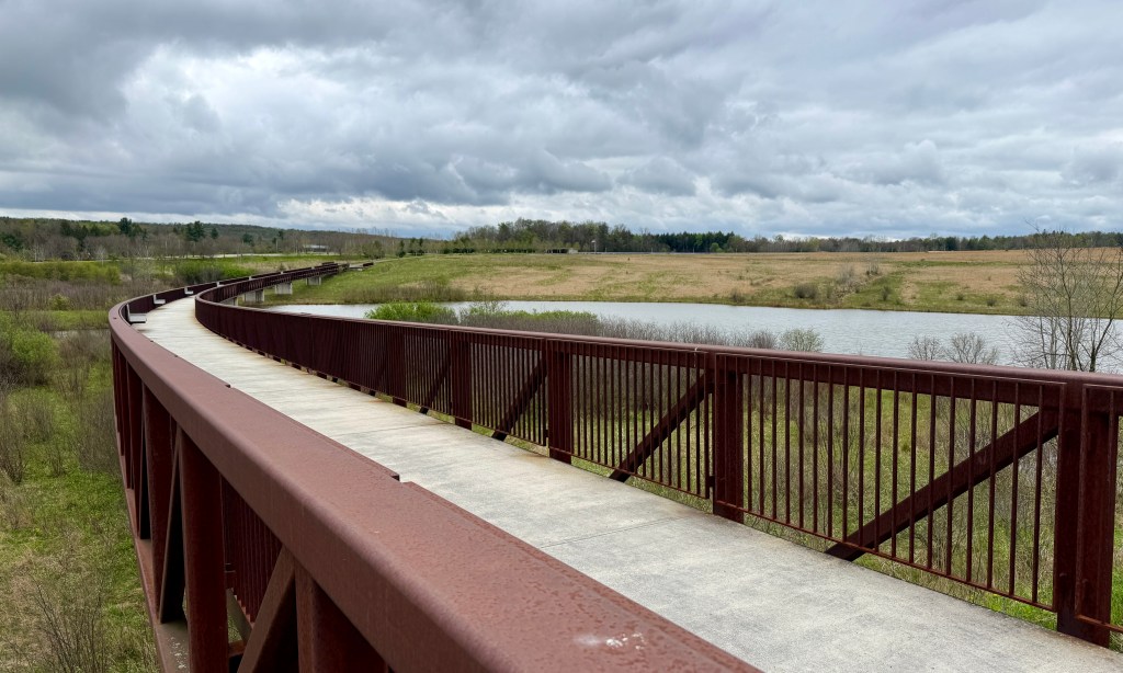 Curved walking path along a river, surrounded by green fields and cloudy skies.