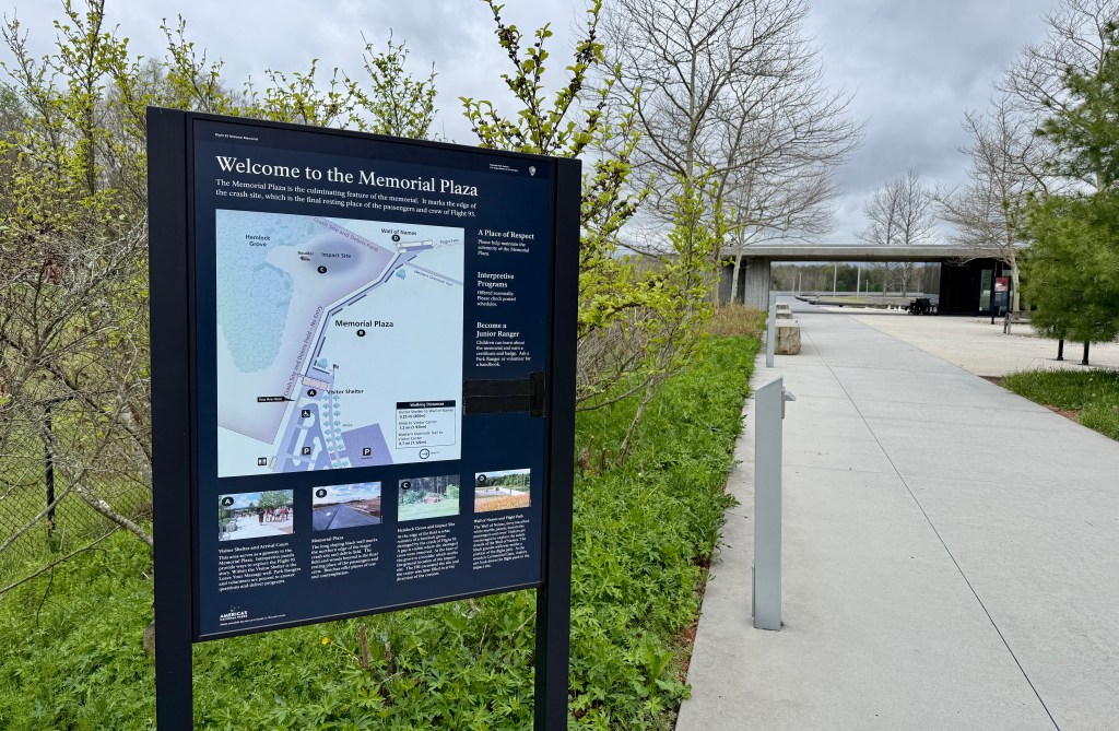 Signage welcoming visitors to Memorial Plaza, with a map and information about the site, surrounded by greenery and a paved walkway.