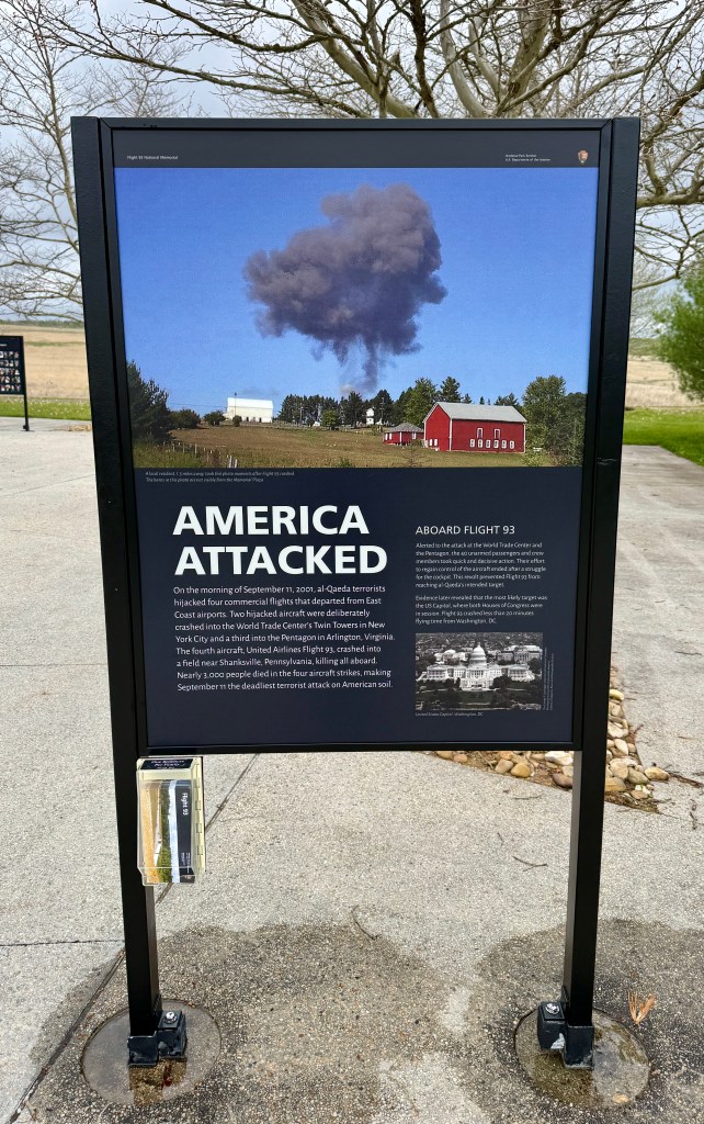 A sign at a memorial site referencing the September 11 attacks, featuring an image of a large smoke cloud in the sky over a rural landscape with farm buildings.