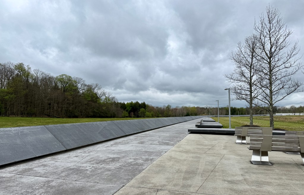 A wide view of a memorial site featuring a long, sloping stone wall, surrounded by grassy areas and trees under a cloudy sky. There are empty benches positioned on a concrete path, inviting contemplation and reflection.