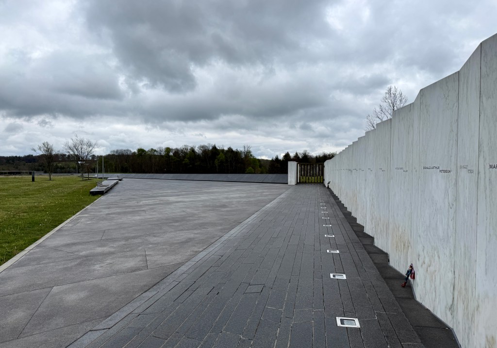 A scenic view of a memorial site featuring a wall inscribed with names, a paved walkway, and a cloudy sky overhead.