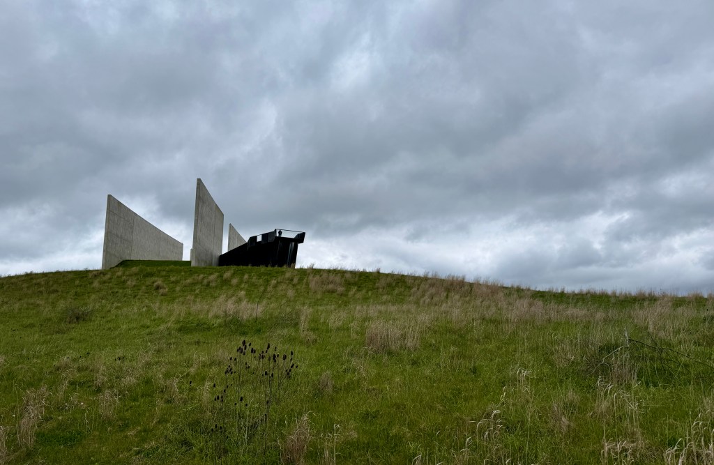 A modern architectural structure made of concrete and metal sits atop a grassy hill, under a cloudy sky.