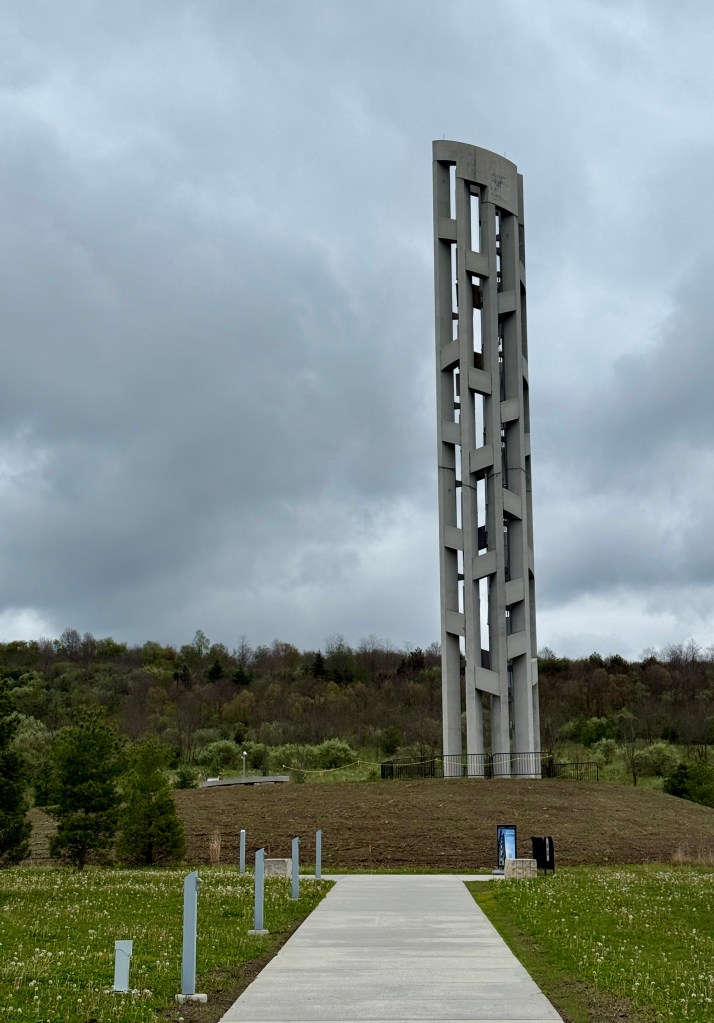 A tall, modern concrete tower standing on a grassy hill, with a walkway leading towards it under a cloudy sky.