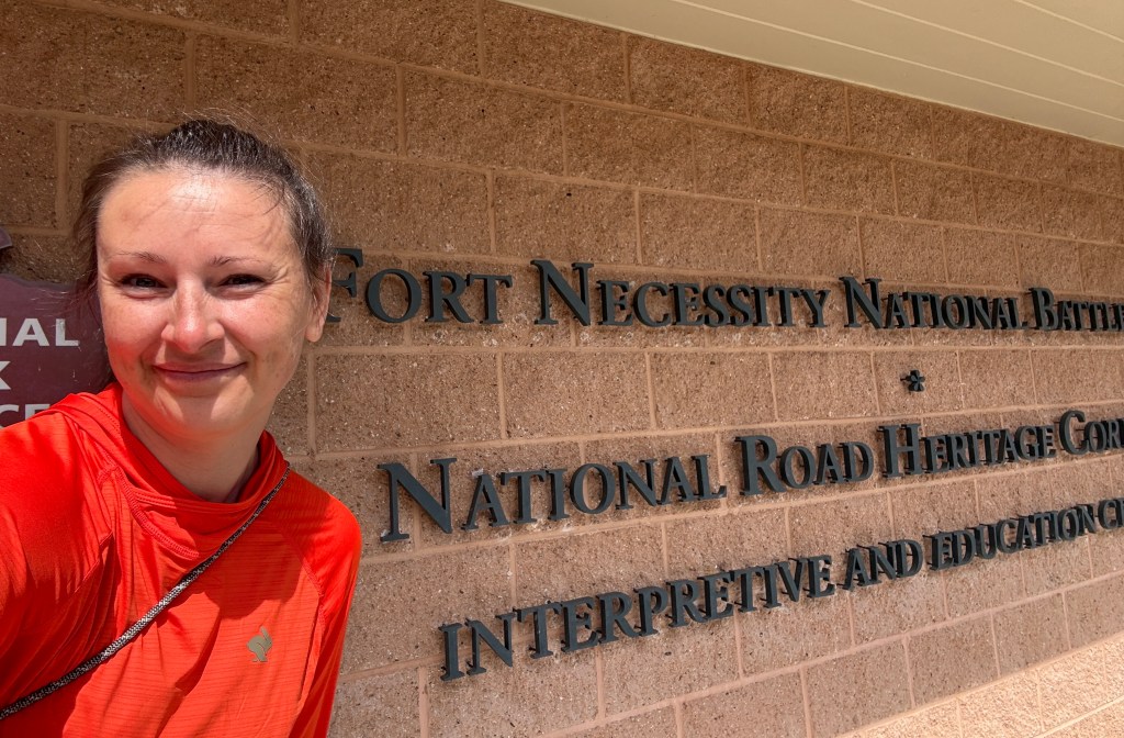 A person smiling in front of the Fort Necessity National Battlefield sign, showcasing the National Road Heritage Corridor.