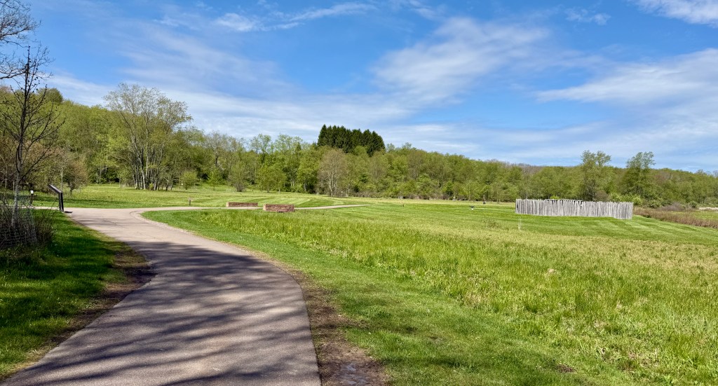 A winding pathway through a lush green park with trees and a cloudy blue sky in the background.