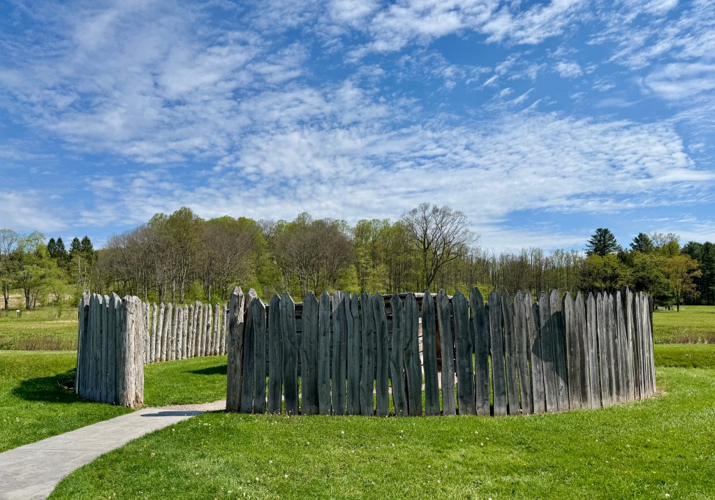 A circular wooden stockade surrounded by green grass and trees under a blue sky with scattered clouds.