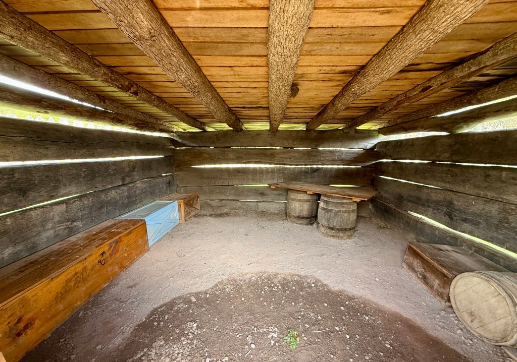 Interior of a rustic wooden shelter featuring log walls, a dirt floor, benches, and barrel-style tables.