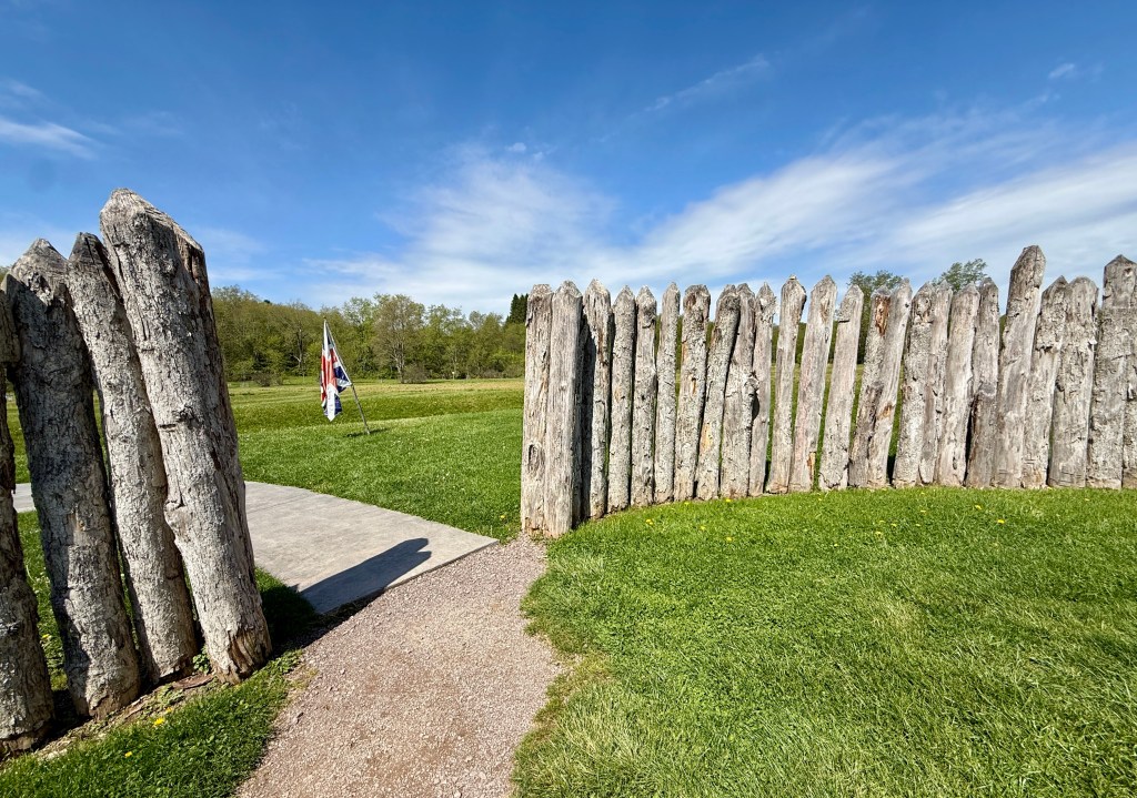An entrance to a grassy field, framed by a wooden post fence with a pathway leading through it, under a blue sky with scattered clouds.