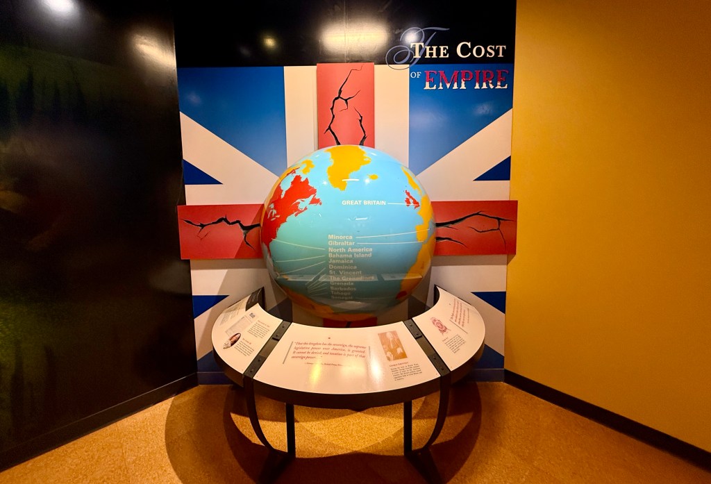 A globe depicting Great Britain with a background featuring the British flag, and text highlighting locations like Minorca, Gibraltar, and the Bahamas, as part of an exhibit titled 'The Cost of Empire'.