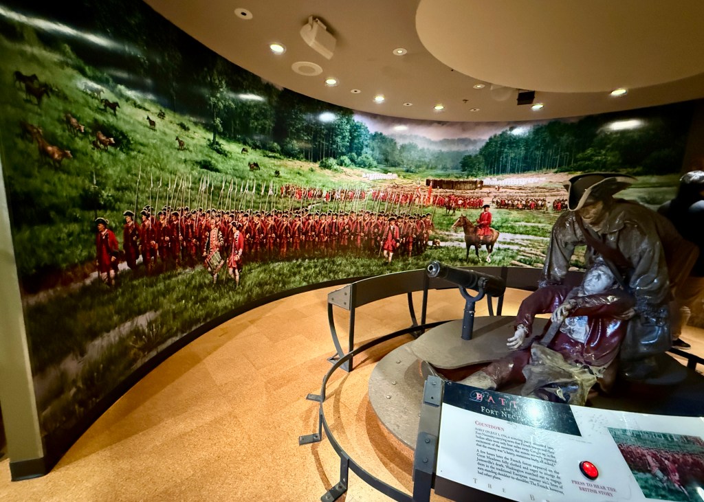 A historical display featuring a mural of soldiers in red coats marching through a green landscape, with a bronze sculpture depicting a military scene in the foreground at a museum.