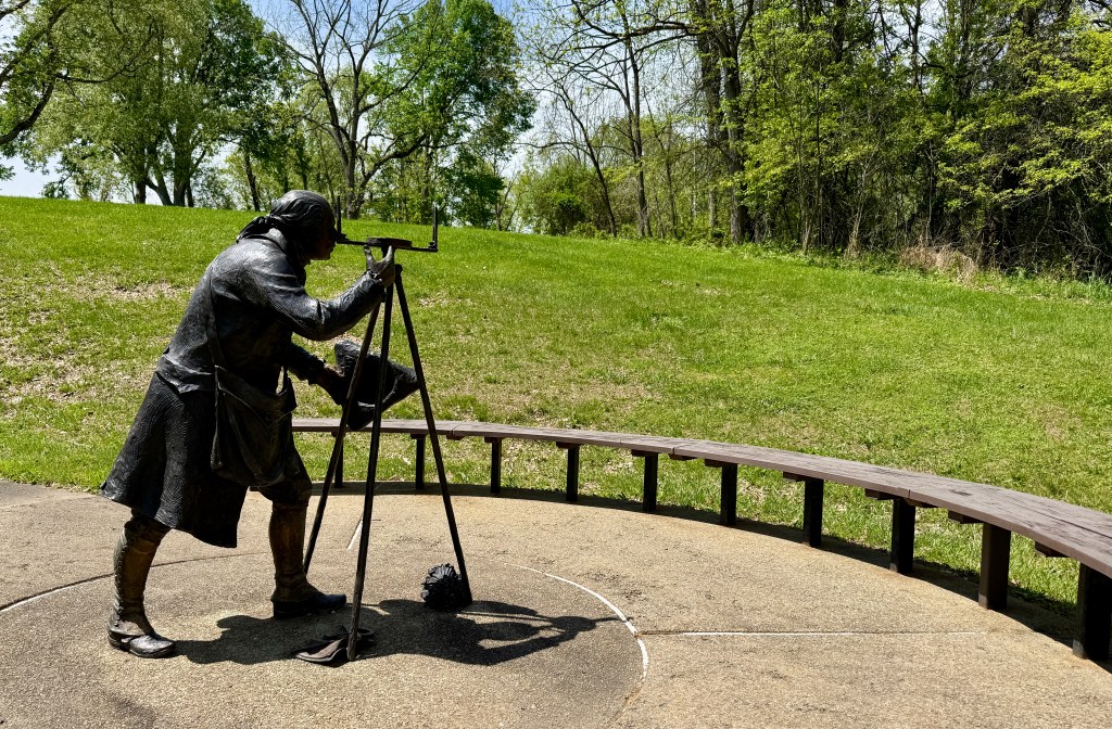 Bronze statue of a man using a telescope on a grassy hill, with trees in the background.