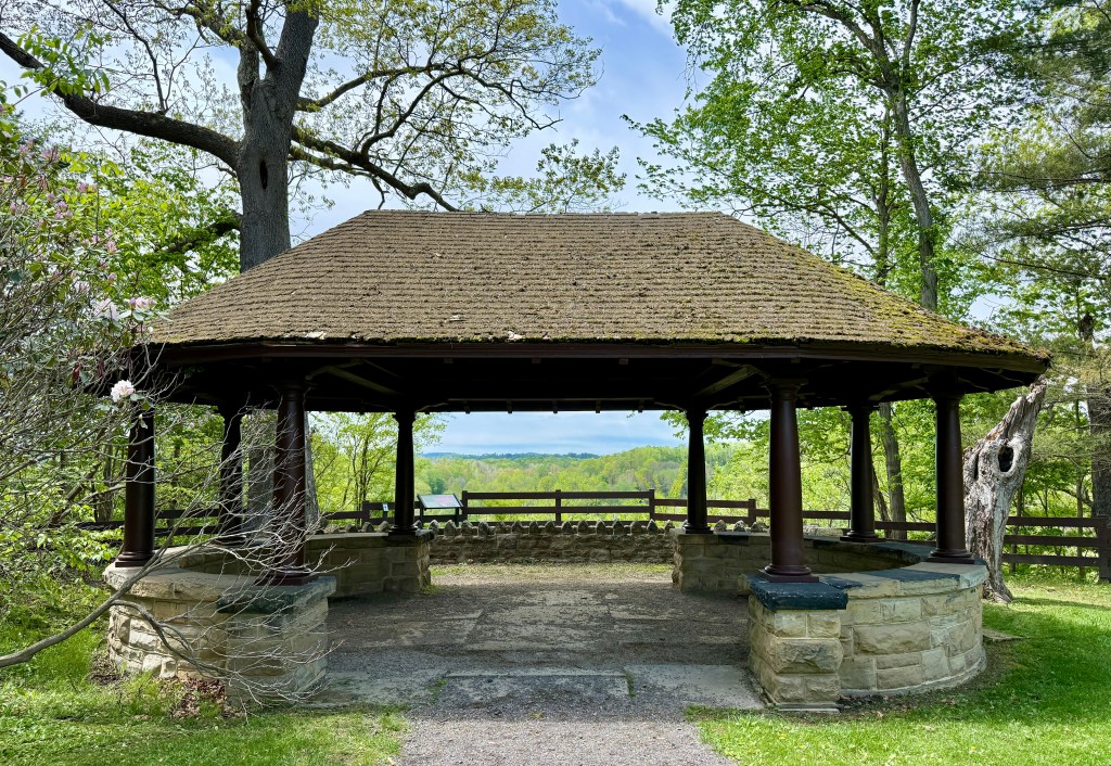 A rustic gazebo with a thatched roof and stone pillars, surrounded by greenery and trees, overlooking a scenic view.
