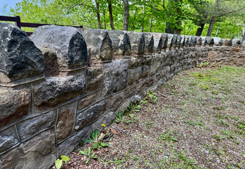 A close-up view of a stone wall with rounded top stones, surrounded by greenery and gravel.