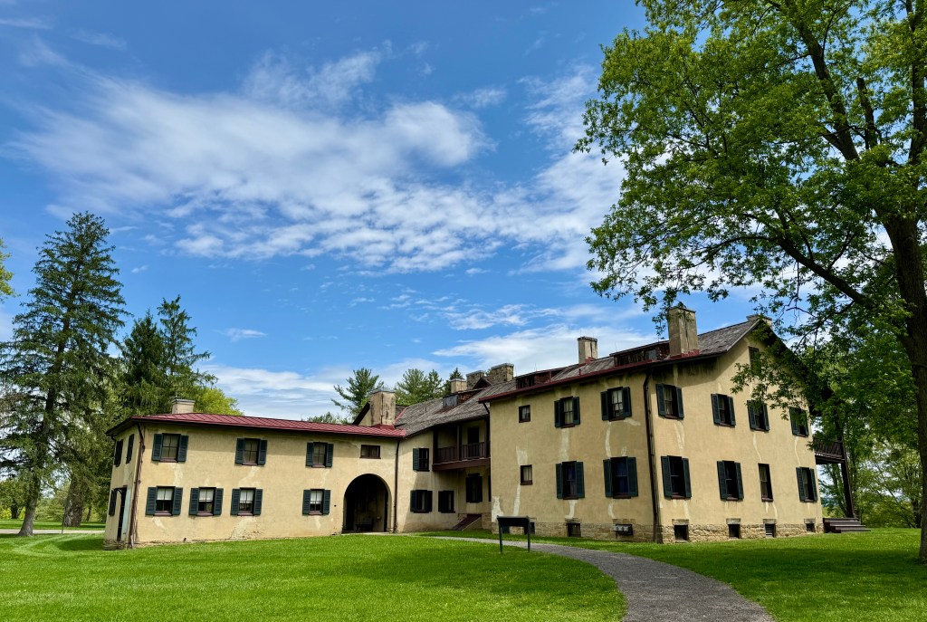 Historic building with a yellow exterior and dark shutters surrounded by green grass and trees under a blue sky.