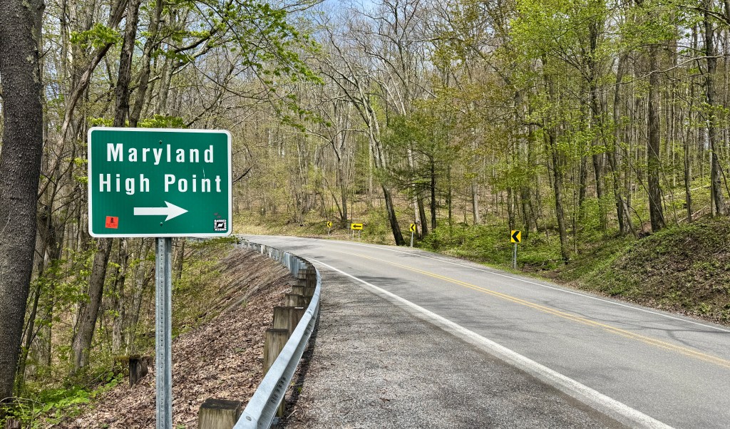A roadside sign indicating the direction to Maryland High Point, with a winding road and greenery surrounding it.