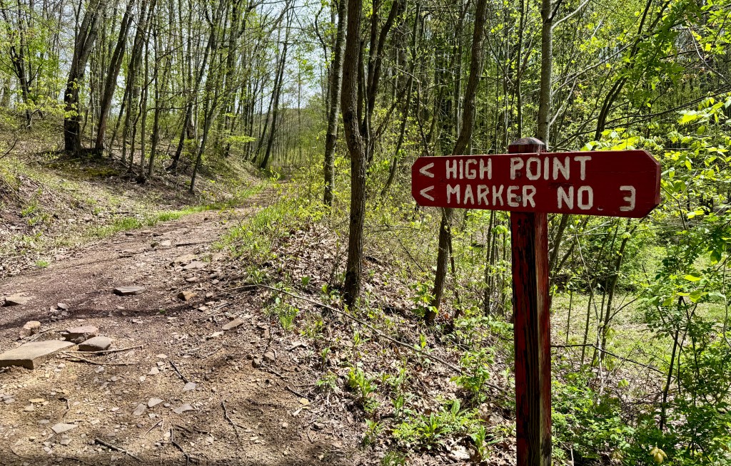 Wooden trail sign indicating directions to 'High Point' and 'Marker No. 3' on a forest path surrounded by green foliage.