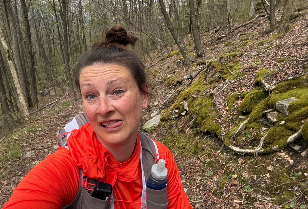 A person hiking on a wooded trail, wearing an orange top and hydration pack, making a funny face.