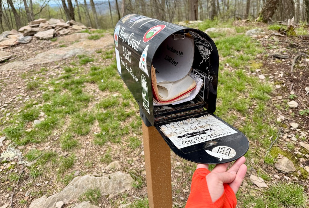 A hand reaching towards an open mailbox on a hiking trail, with a sign indicating the location and instructions for visitors.