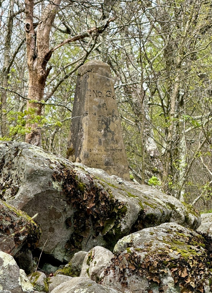 A weathered monument stone engraved with the number 'No. 3' and names, surrounded by rocky terrain and bare trees in a forest setting.