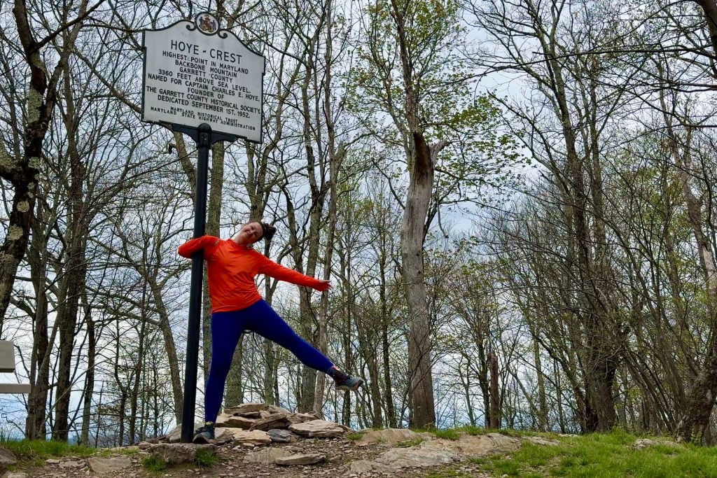Person posing joyfully next to a Hoye Crest sign marking the highest point in Maryland, surrounded by trees and rocky terrain.