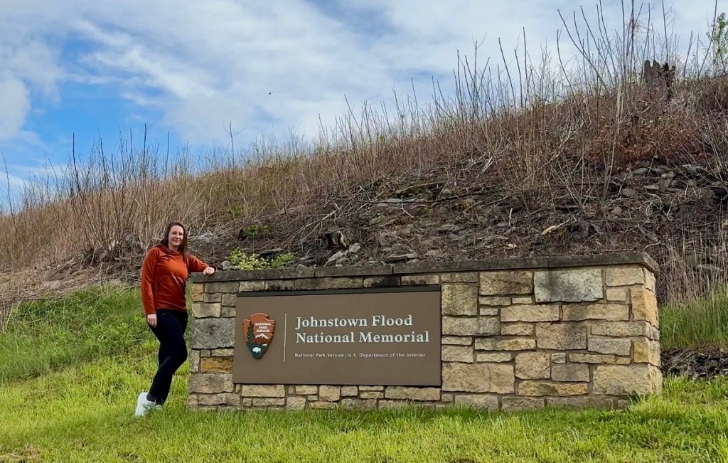 A woman in an orange top and black pants stands next to a stone sign that reads 'Johnstown Flood National Memorial' with a blue sky and grassy area in the background.