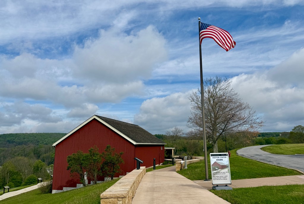 A scenic view of a red barn alongside a pathway, with an American flag waving in the foreground and green hills in the background, under a cloudy sky.