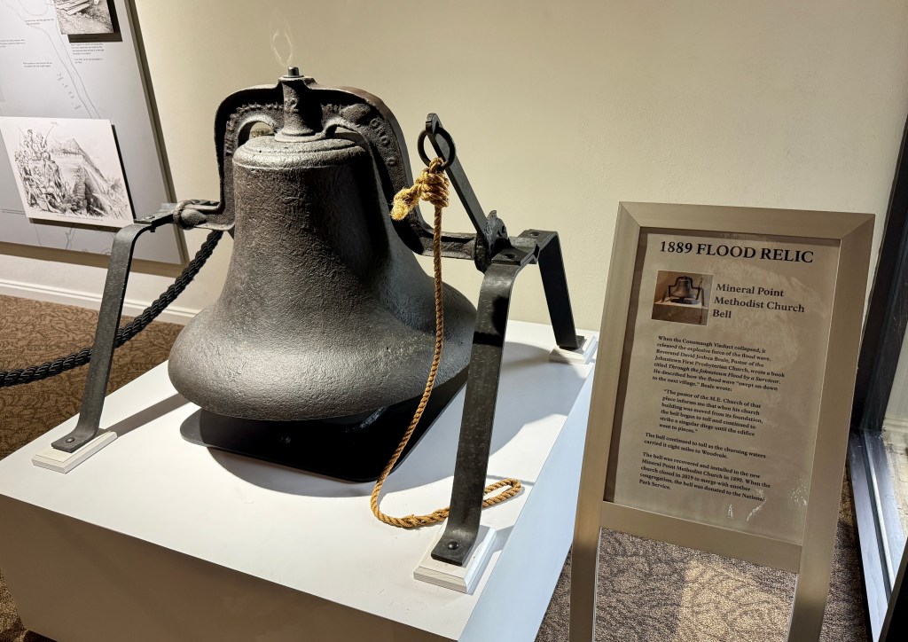 A historical bell from the Mineral Point Methodist Church displayed on a stand, with a sign detailing its significance as a relic from the 1889 flood.