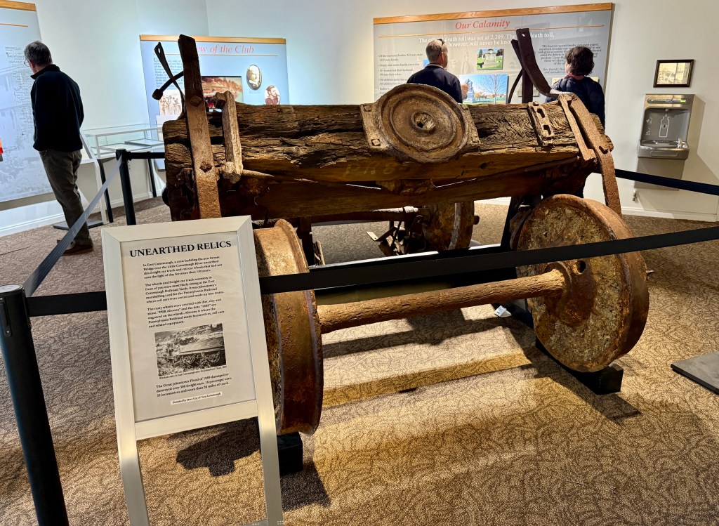 A historic artifact displayed in a museum, featuring a rusted old wagon or cart with wooden wheels and metal components, surrounded by barriers. Visitors observe the exhibit while informational panels are visible in the background.