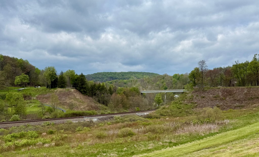 A scenic view of rolling hills under a cloudy sky, with a railway track in the foreground and a bridge in the background.