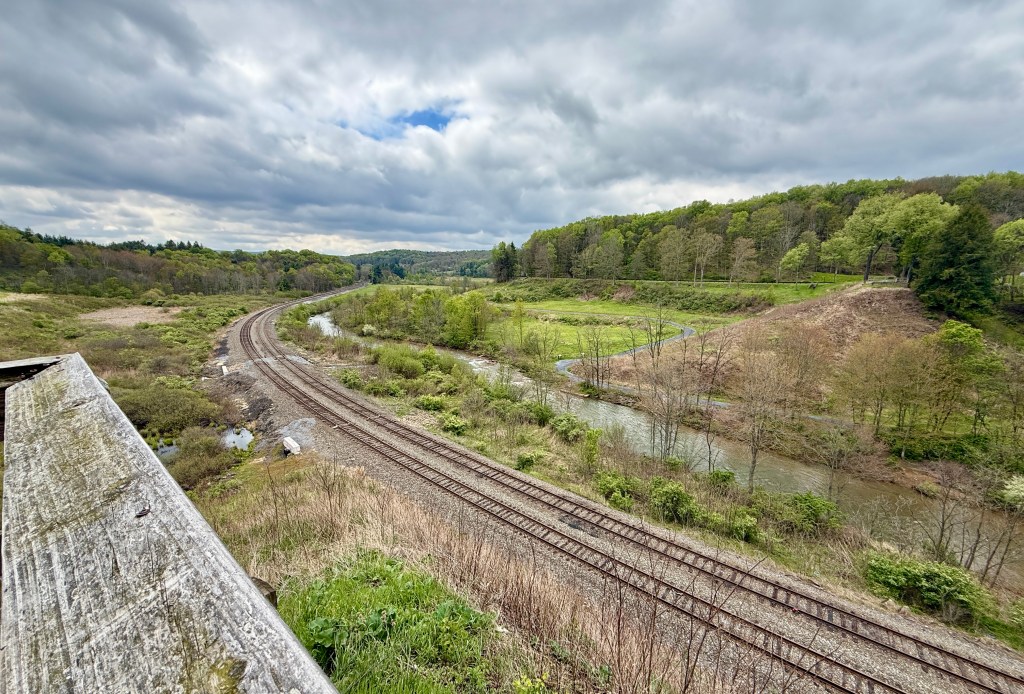 A scenic view of a winding railway track through a green landscape, with a river flowing alongside and cloudy skies above.