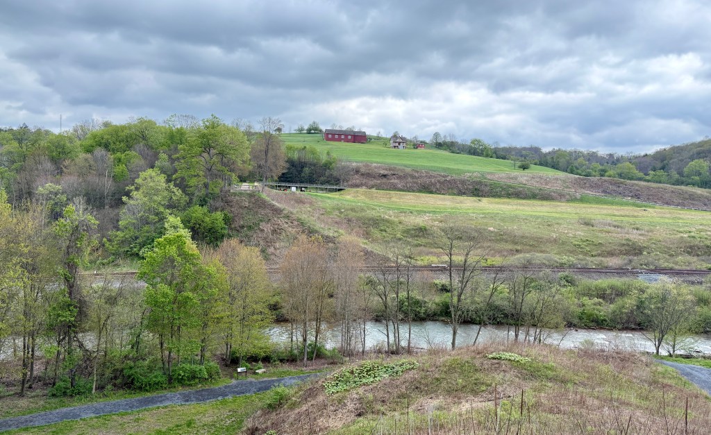 A landscape view featuring a meandering river, green hills, and trees, with cloudy skies overhead and red barns in the distance.