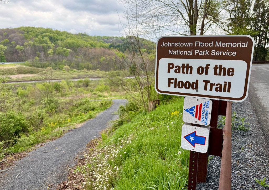 Sign for the Path of the Flood Trail at Johnstown Flood Memorial National Park, showing a gravel path leading through lush greenery.