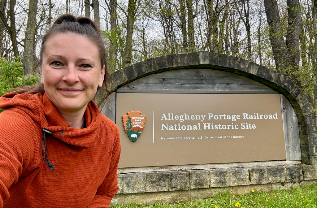 A person in an orange hoodie smiling in front of the Allegheny Portage Railroad National Historic Site sign, surrounded by trees.