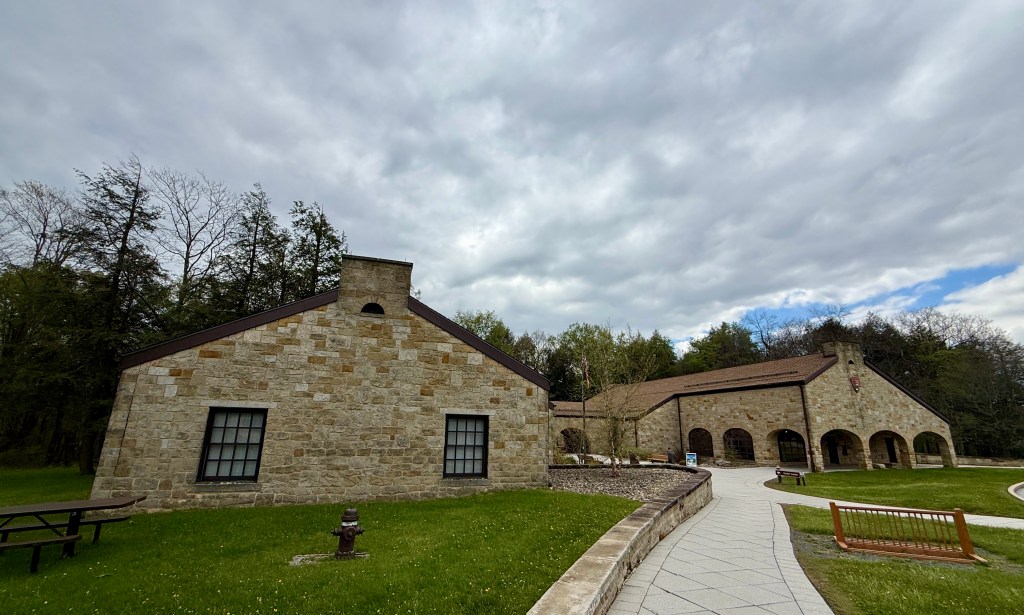 Exterior view of a stone building with a curved pathway, surrounded by green grass and trees under a cloudy sky.